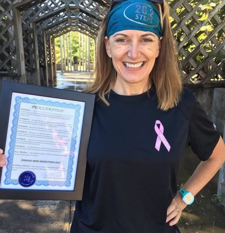 Denise Ibsen Cole smiling outdoors, wearing a 26.2 Steps headband and holding a framed City of Omaha Mini Marathon Day proclamation